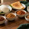 Four small bowls of podi beside idli, dosa and rice on a wooden table